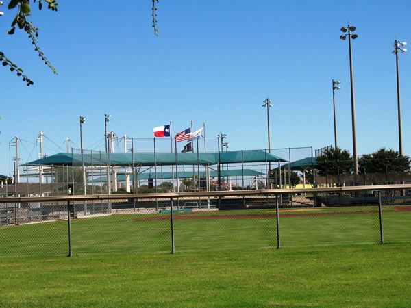View of the Five Star Complex featuring multiple baseball or softball fields with green shade structures over the bleachers. The scene includes tall field lighting poles, a well-maintained grassy area, and three flags flying—Texas, the U.S., and a third flag—against a clear blue sky.