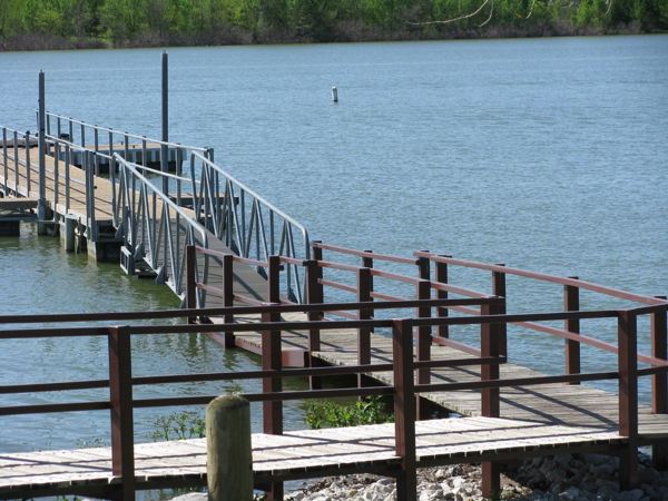 Courtesy Dock at Stewart Creek Park extending into the lake with a sloped metal gangway and wooden walkway, surrounded by calm water and bordered by a rocky shoreline and trees in the background. Ideal for loading and unloading boats.