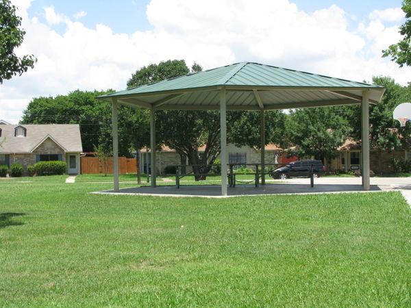 Pavilion at Carr-Chapman Park featuring a green metal roof supported by open columns, situated on a concrete pad surrounded by a well-maintained grassy lawn, with residential homes and trees in the background. Ideal for shade and gatherings.