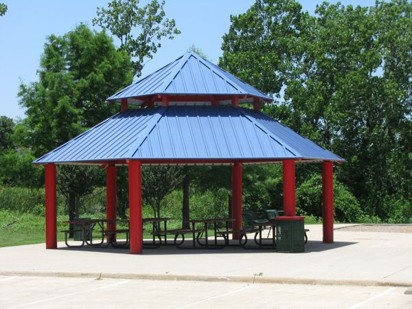 Pavilion at Ridgepointe Park featuring a vibrant blue double-tiered metal roof supported by bold red columns, with multiple picnic tables underneath on a concrete pad, surrounded by lush greenery.