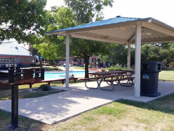 Pavilion at the Aquatic Park with a blue metal roof and open sides, covering picnic tables on a concrete pad. Nearby are barbecue grills, a trash can, and a splash pad play area surrounded by trees and park amenities.