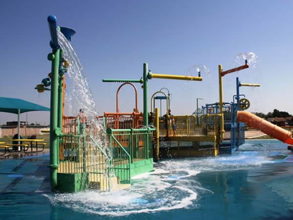 Colorful Splash Zone at the Aquatic Park featuring interactive water play structures, dumping buckets, sprayers, and slides surrounded by a shallow pool area under a clear blue sky.