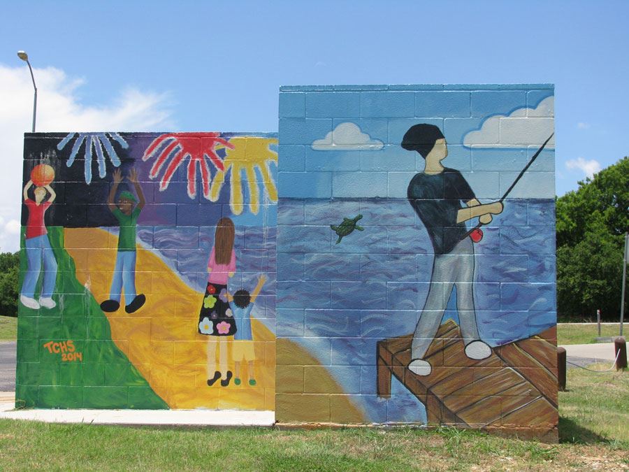 Colorful mural at Stewart Creek Park painted on a block wall, depicting children playing under fireworks and a woman with two kids walking along a path on one side, and a person fishing from a wooden dock over a lake with a turtle swimming nearby on the other side. The mural is signed “TCHS 2014” and set against a sunny blue sky with grassy surroundings.