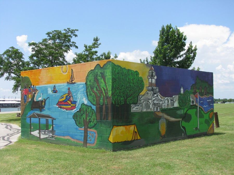 Mural at Stewart Creek Park dumpster container painted on a cornered block wall, illustrating vibrant outdoor scenes. One side shows lake activities including boating, jet skiing, and fishing, along with a shaded picnic area. The adjacent side features a forested area, tents, a campfire, a historic-style building, and a golf course with a flagstick near a water hazard. The background transitions from a warm sunset to a cool twilight sky, symbolizing a full day of recreational fun.