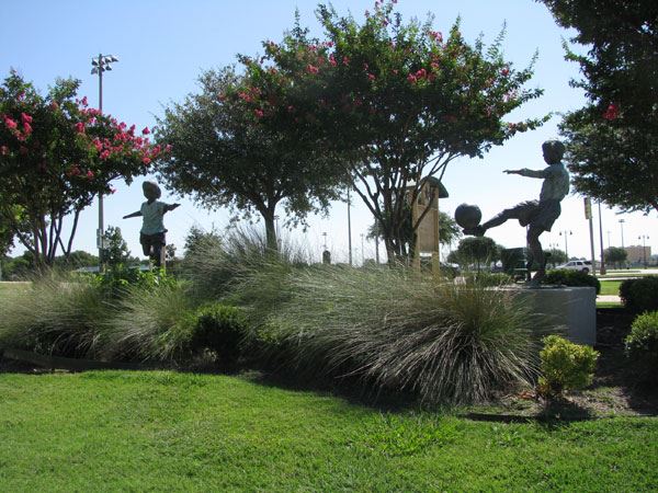 Bronze sculptures at Five Star Complex depicting children playing. The statues show lifelike figures of kids engaged in joyful outdoor activities, with one child poised to kick a soccer ball. The sculptures are surrounded by manicured landscaping, ornamental grasses, and flowering crepe myrtle trees. Sports field lights are visible in the background, indicating the area’s recreational use. The scene captures a vibrant and welcoming community atmosphere.