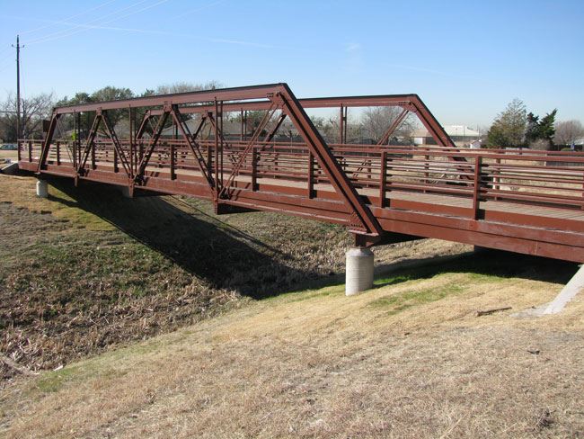 Historic metal truss bridge at Greenway Park, repurposed for pedestrian use. The rust-colored bridge spans a dry creek bed and is supported by concrete piers. It features a wooden deck and railing, with a distinctive triangular truss design. The bridge is a preserved structure reflecting early 20th-century engineering and serves as a functional and aesthetic element of the park's trail system. Leafless trees and dry grass indicate a winter season, with clear blue skies overhead.