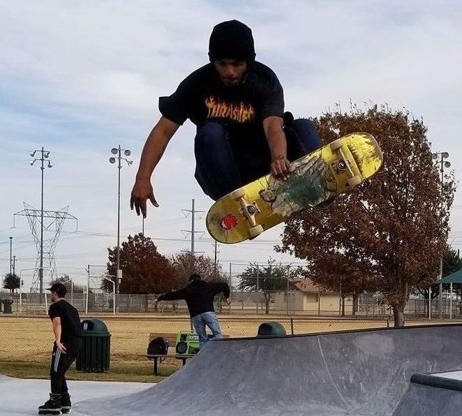 A skateboarder wearing a black hoodie and Thrasher t-shirt performs an aerial trick at a concrete skatepark, captured mid-jump above a ramp with a yellow skateboard. In the background, other skaters and trees with fall foliage can be seen, along with a fenced sports field and tall light poles, suggesting a multi-use recreational park area.