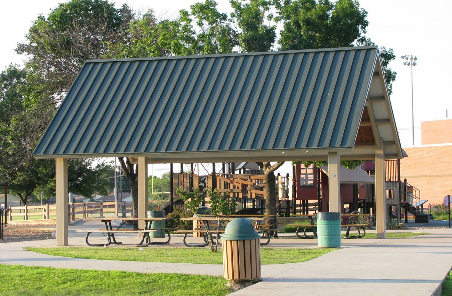  Kids Colony Pavilion featuring a large metal-roofed shelter with picnic tables underneath. The pavilion is situated in a park setting, adjacent to a wooden playground structure with ramps and slides. Surrounding the area are green trees, walking paths, and trash bins, creating a family-friendly environment ideal for gatherings and outdoor play.