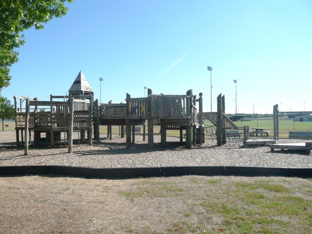Old Playground at Kids Colony featuring a large, elevated wooden play structure with multiple platforms, bridges, and slides. The playground is set on a mulch surface and surrounded by open grassy fields and sports lighting in the background, indicating nearby athletic facilities. The design has a rustic, fort-like appearance, popular in earlier playground styles.