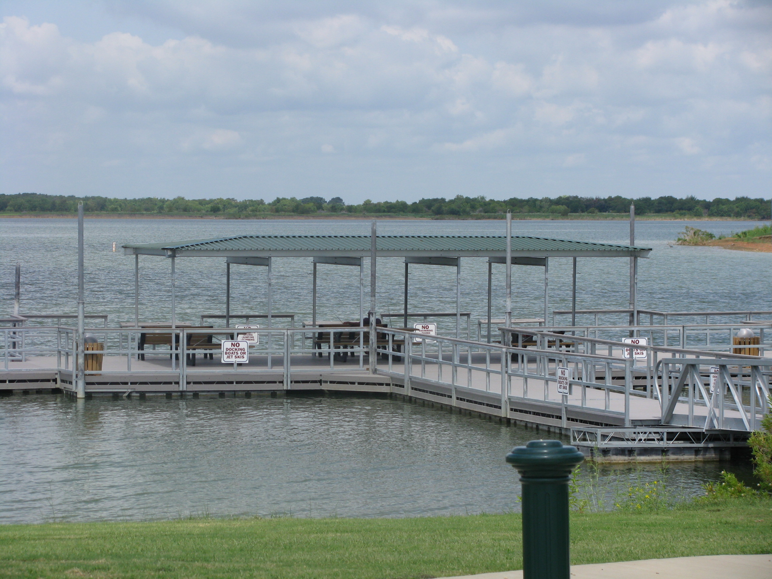 Fishing Pier at Stewart Creek Park extending over a calm lake, featuring a covered section with benches for visitors. The pier has metal railings and multiple signs indicating "No Swimming" and fishing regulations. The surrounding landscape includes grassy banks, water, and a tree-lined shoreline in the background under a partly cloudy sky.