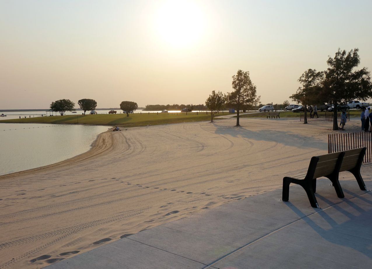 Swim Beach at Stewart Creek Park during sunset, featuring a sandy shoreline leading to a calm body of water. A few benches are placed along a concrete path near the beach. Trees and parked cars line the edge of the park, with people enjoying the scenic view and open space. The setting sun casts long shadows and a warm golden hue over the scene.